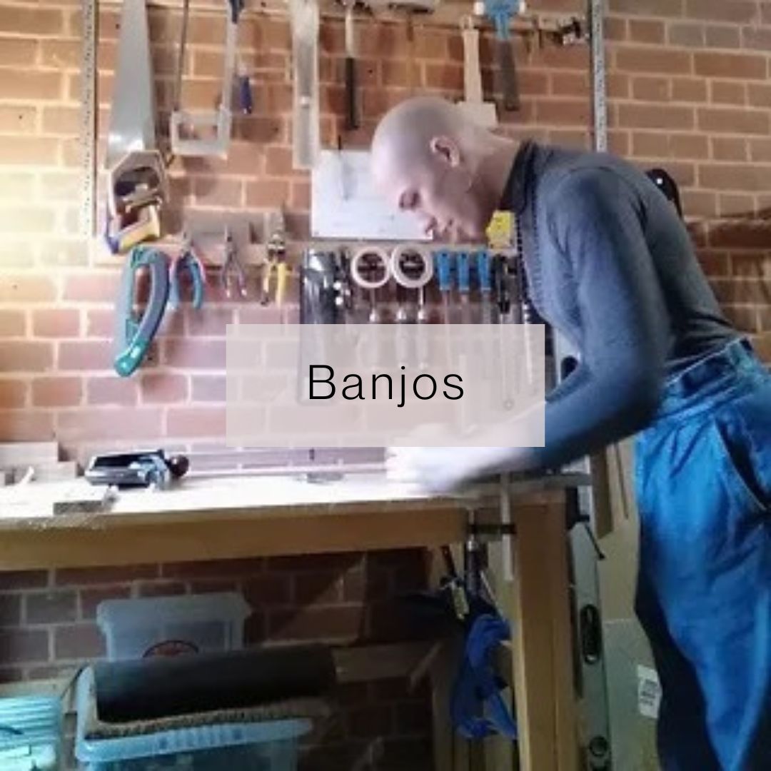 Jacken stands at a work bench with tools on the wall behind. She is wearing blue jeans and a grey top, and is working on a banjo.