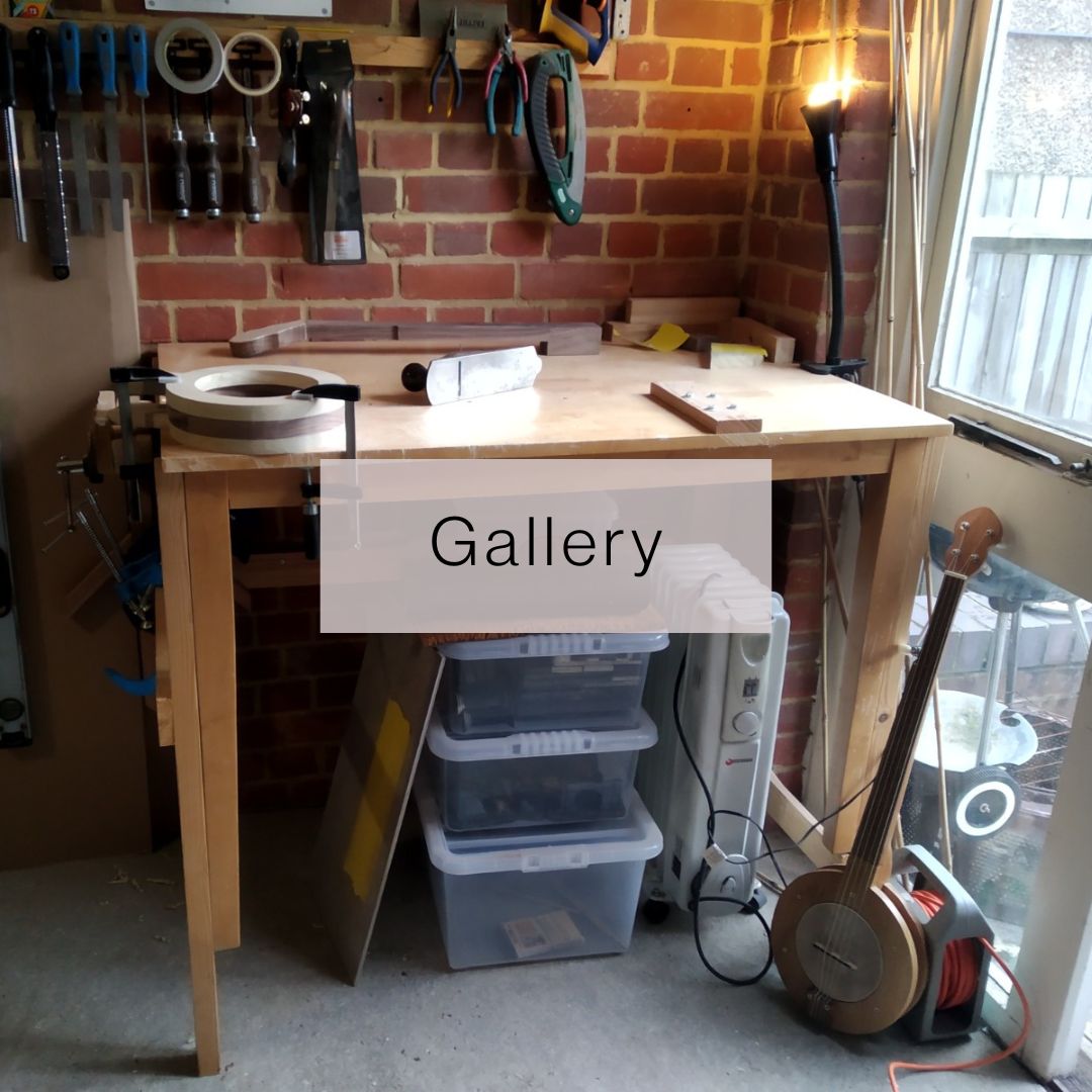 A workbench stands against a brick wall, with various tools hanging from it or stored underneath it. One banjo is in-progress on the bench, another stands to one side.