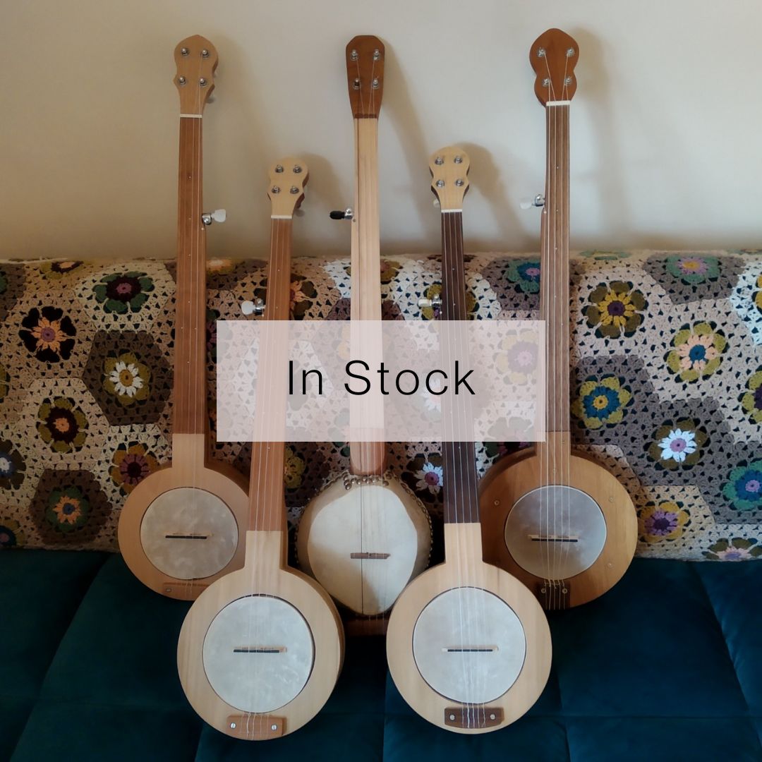 Five banjos made from a variety of woods stand against a crocheted quilt. They are all fretless, four are mountain banjos and one is a gourd banjo.